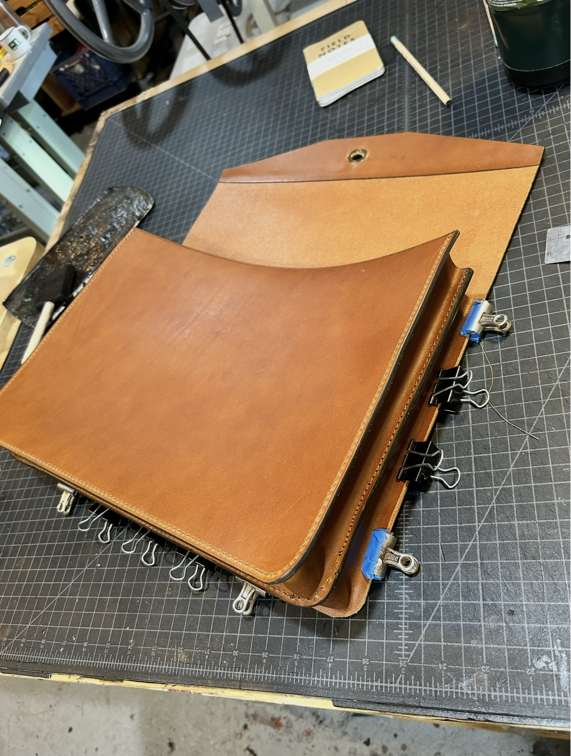 A leather portfolio or binder in process of assembly on a worktable, with binder clips attached, along with a piece of leather, a roller, a small notebook, and a pen.