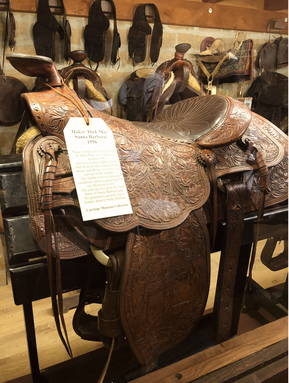 A detailed leather saddle with floral carvings, displayed in a museum setting. A tag hanging from the saddle indicates it was made in Santa Barbara in 1956 by Dick May, and it is part of the Carriage Museum Collection.