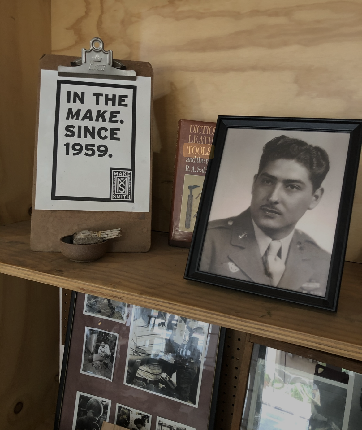 A framed black-and-white portrait photo of a man in a military uniform, a book titled 'Dictionary of Leather Tools,' and a wooden sign with text 'In the Make. Since 1959.'