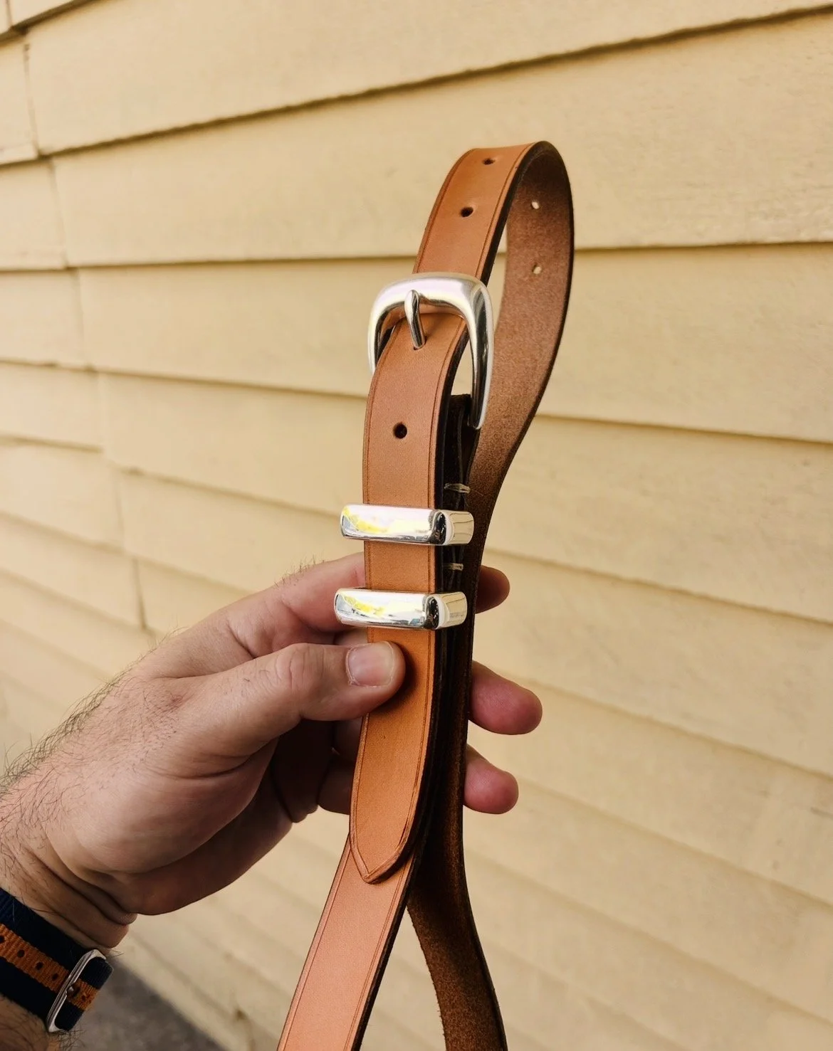 A person's hand holding a tan leather belt with silver hardware against a yellow wooden wall.