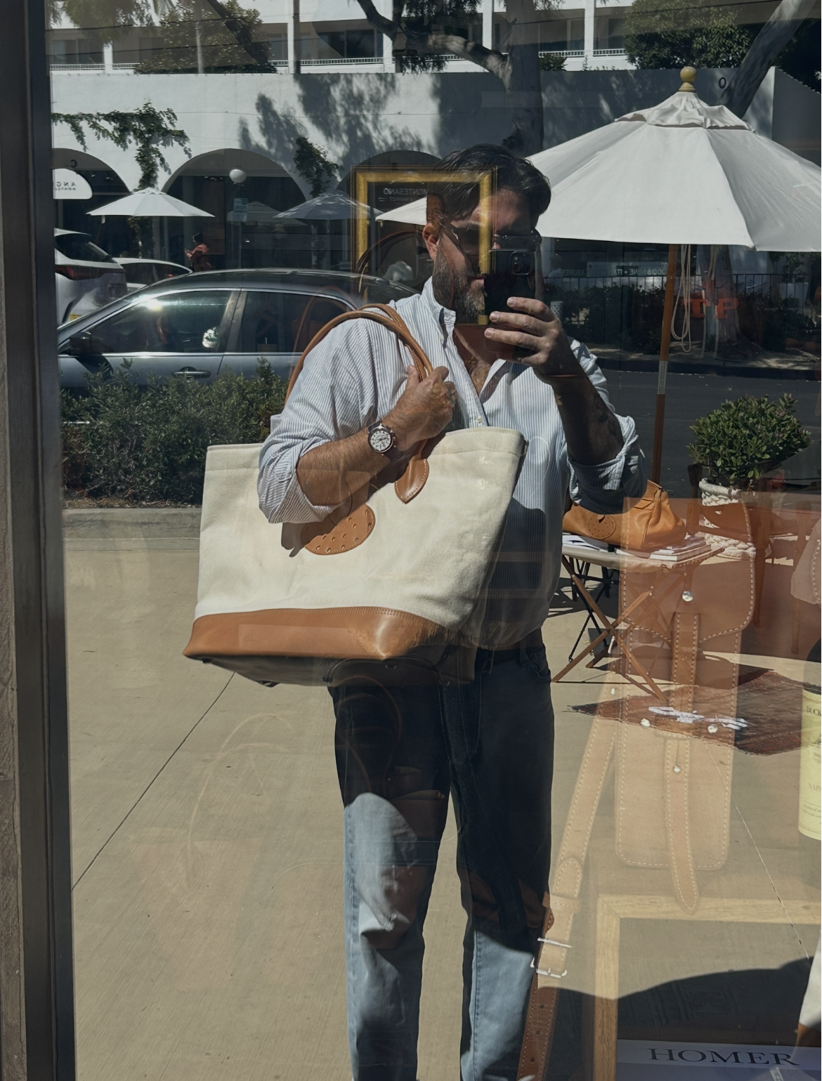 Person taking a photo of their reflection in a storefront window, holding a large beige and brown tote bag, wearing a white and blue striped shirt, sunglasses, and a watch, with outdoor patio umbrellas and parked cars in the background.