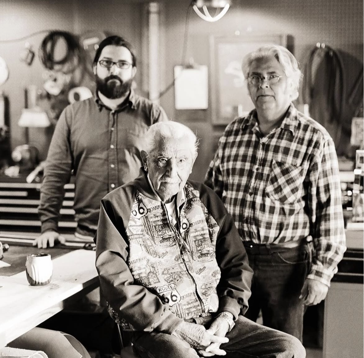Three men and one elderly woman in a workshop or garage setting, with tools and equipment in the background. The woman is seated in front, wearing a jacket with garage-themed prints, and the men are standing behind her, one in a plaid shirt and glass