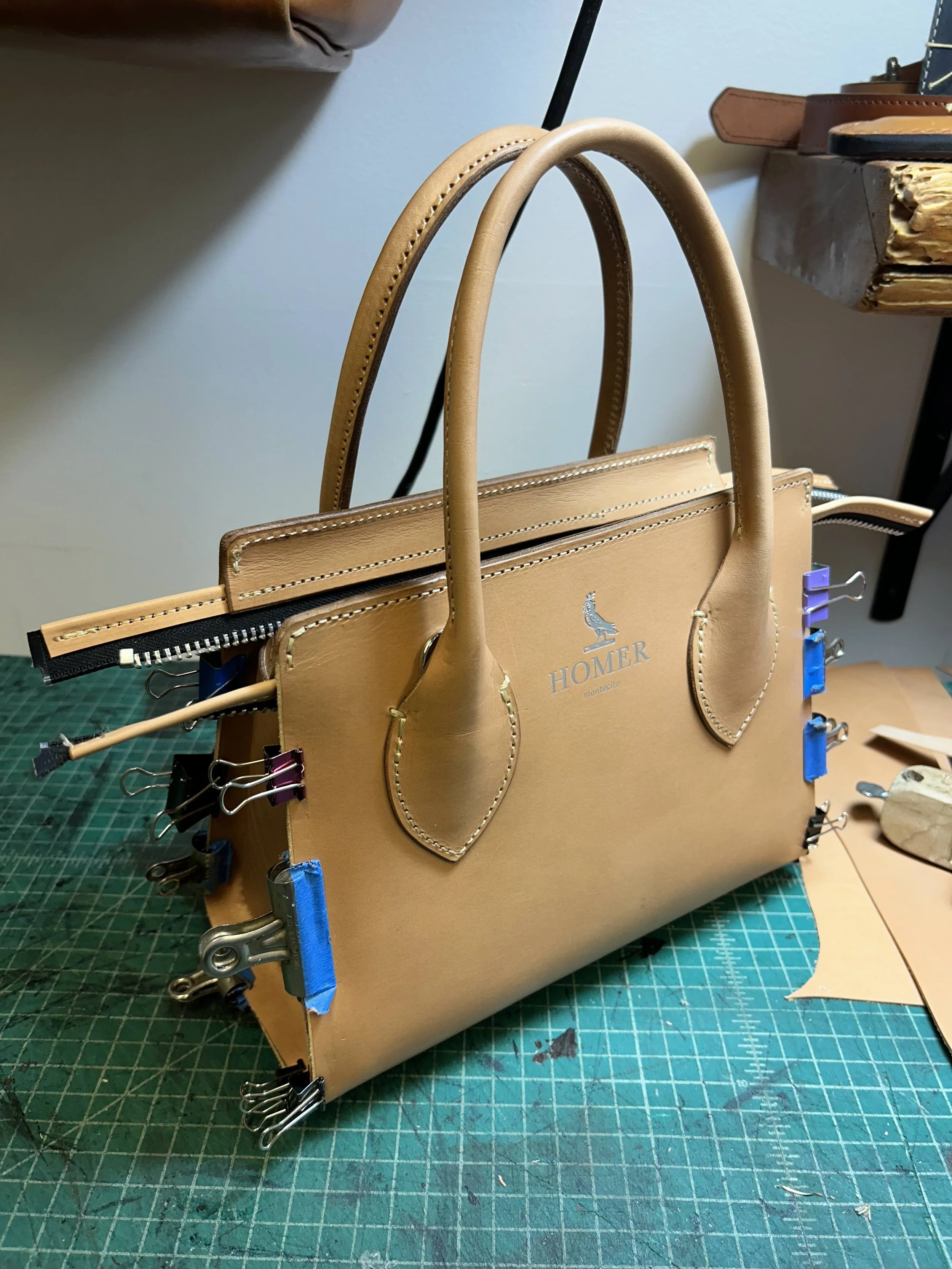 Handcrafted tan leather handbag on a green cutting mat with clips and tools, with the brand name 'HOMER' and a bird logo visible on the front.