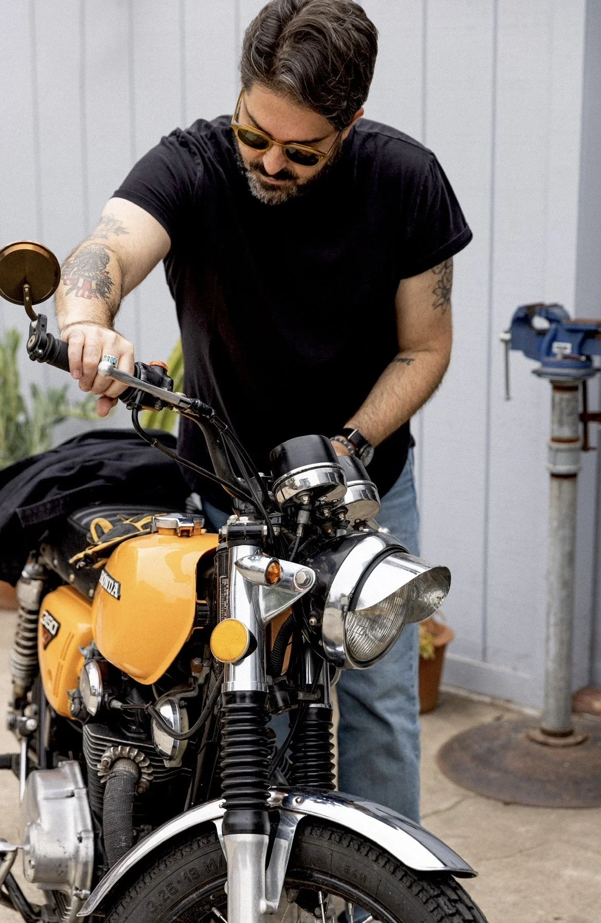 Man with sunglasses and tattoos working on a yellow vintage motorcycle indoors.