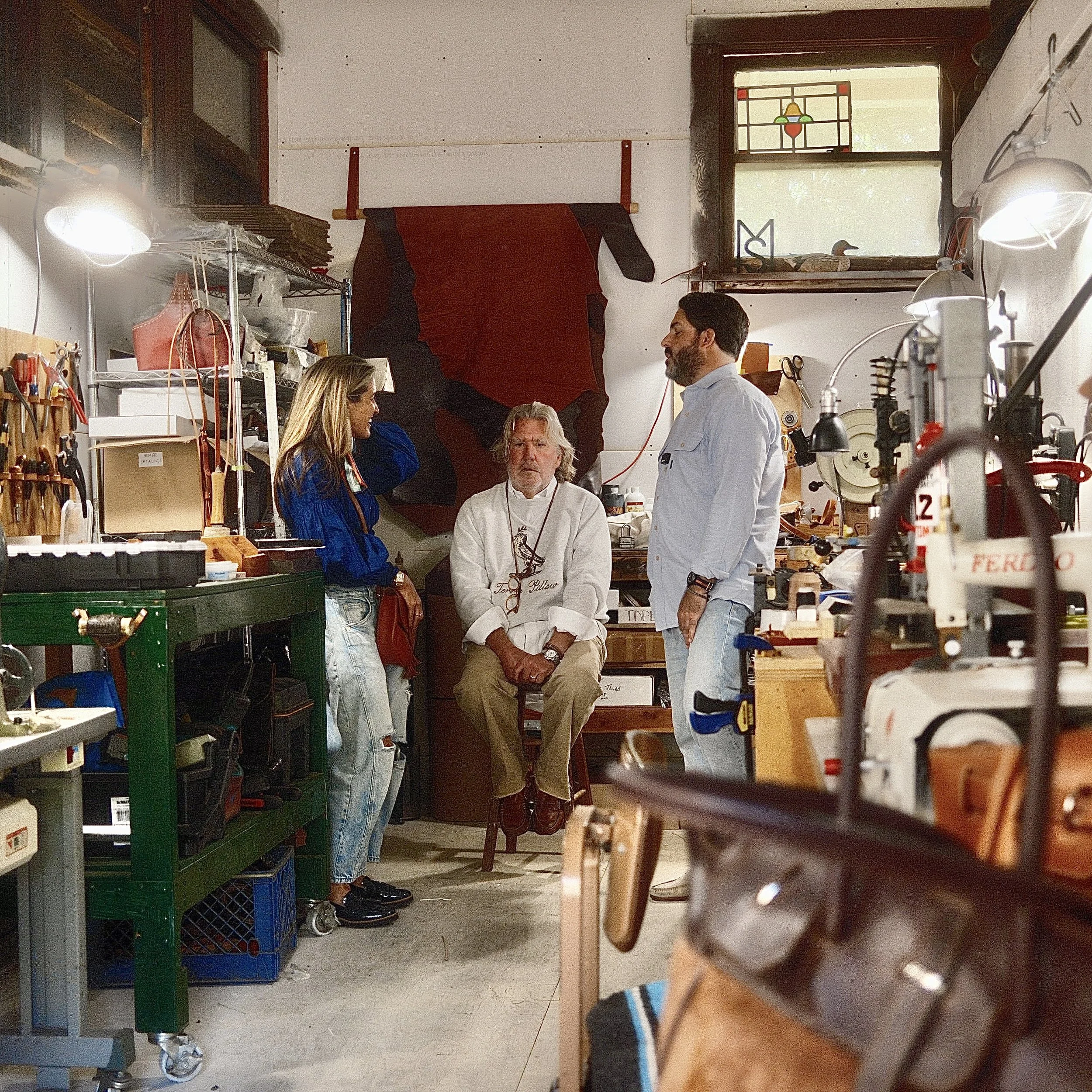 Four people in a workshop engaged in conversation. One older man sits on a stool, while a woman and a man stand on either side of him. Another woman stands nearby, listening. The workshop has shelves with tools and materials, and a window with staine