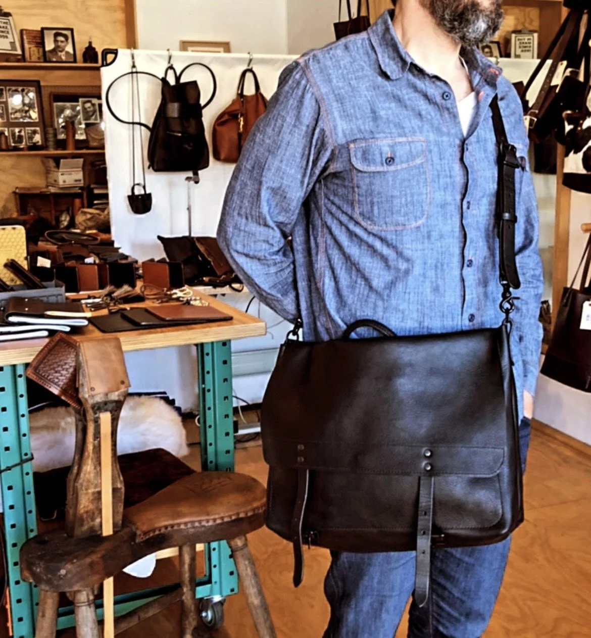 A man wearing a denim shirt with a leather messenger bag hanging from his shoulder stands inside a store with various handbags and wallets displayed on shelves and tables in the background.