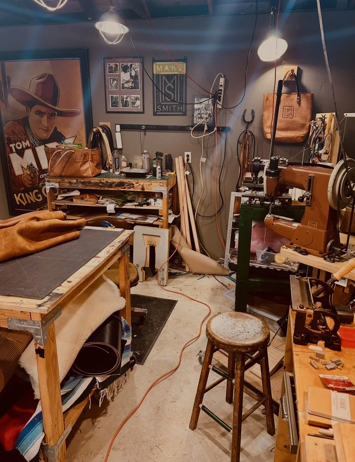 A cluttered workshop with various tools, materials, and artwork. Features a wood table, a sewing machine, a stool, and framed pictures on the wall, including a large poster of a woman wearing a hat.