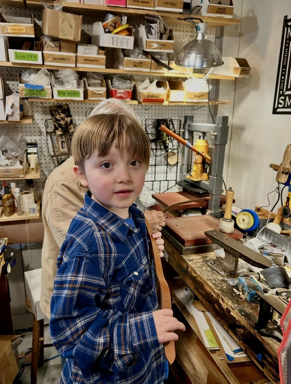 Young boy in a plaid shirt holding a leather strap in a workshop with woodworking tools, shelves with boxes, and a pressed flower among other supplies.