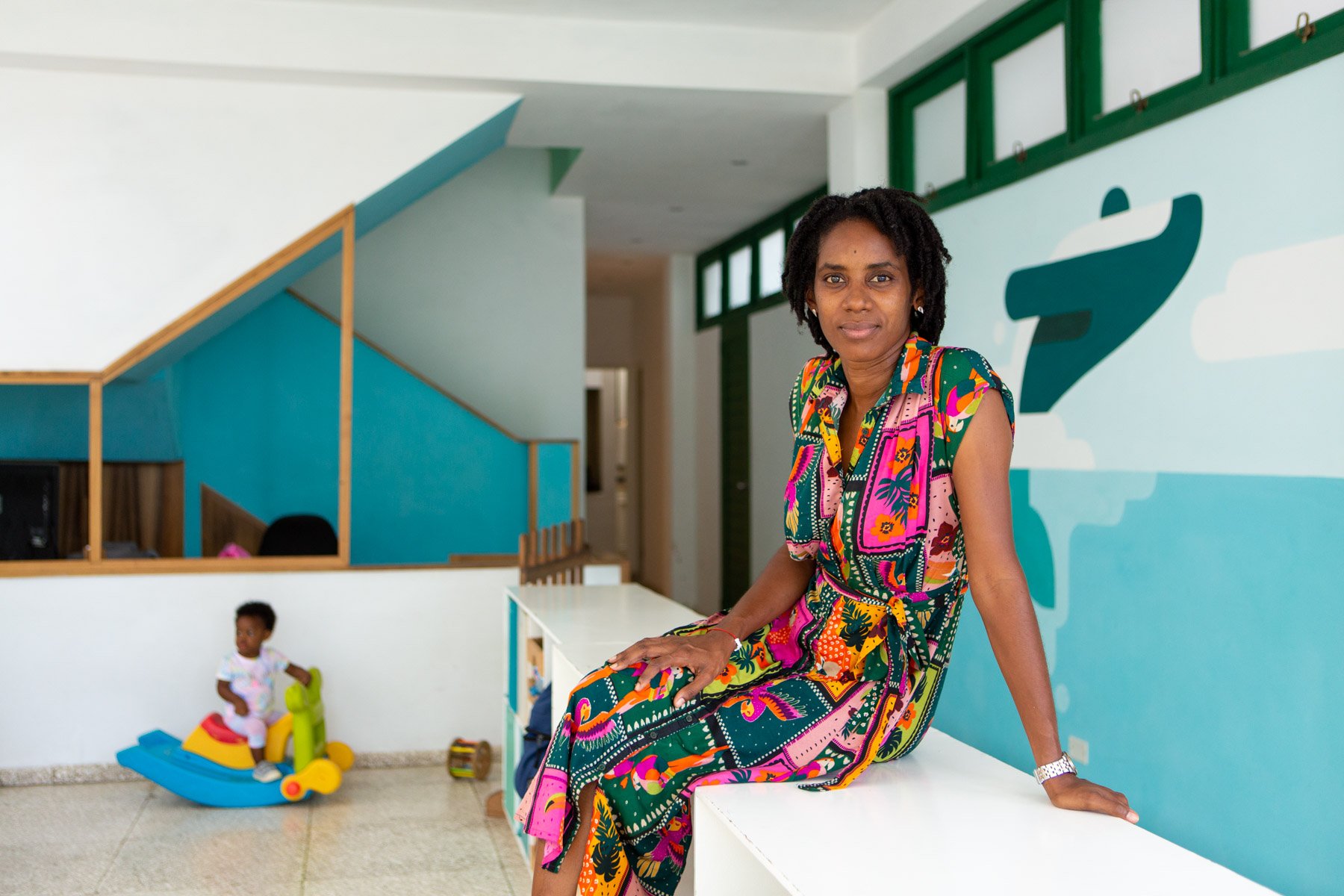 Woman wearing a colorful dress sitting on a white ledge inside a modern building with children playing in the background.