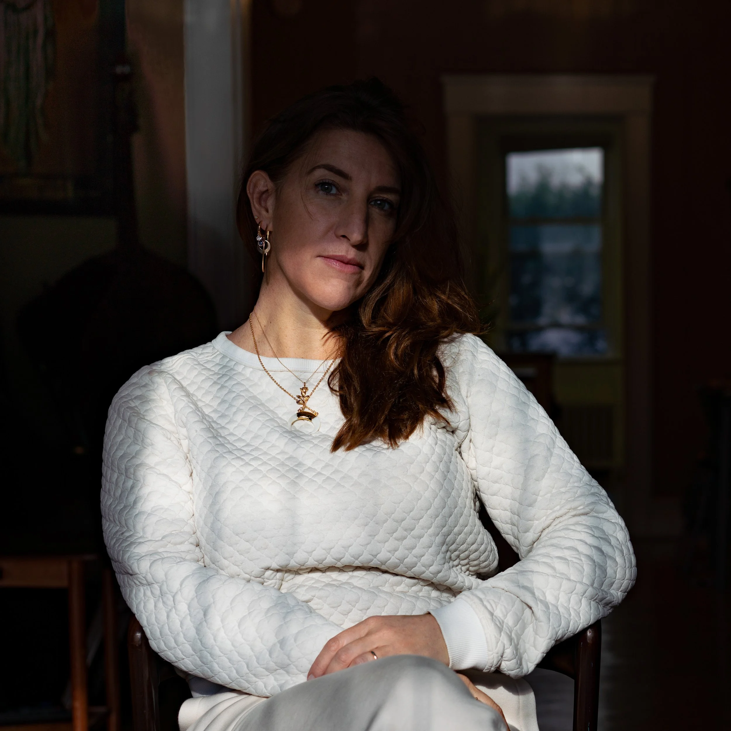 A woman with long brown hair, wearing a white quilted sweater and layered gold necklaces, sitting indoors with a neutral expression, in front of a window showing a cloudy sky.