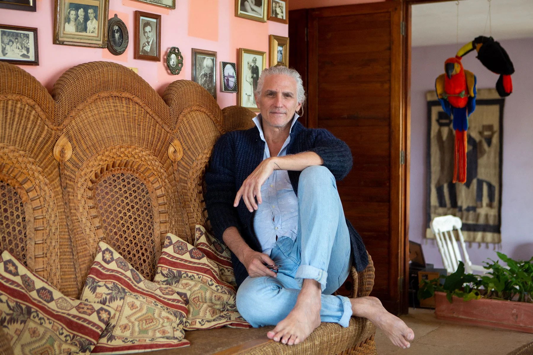 A man sitting on a wicker couch in a cozy living room, surrounded by vintage photos and artwork on the wall, with a colorful parrot hanging in the background.