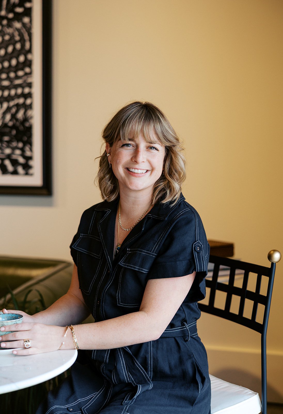 Full smile photo of Laura Rudicle indoors in dark blue dress sitting at a cafe table
