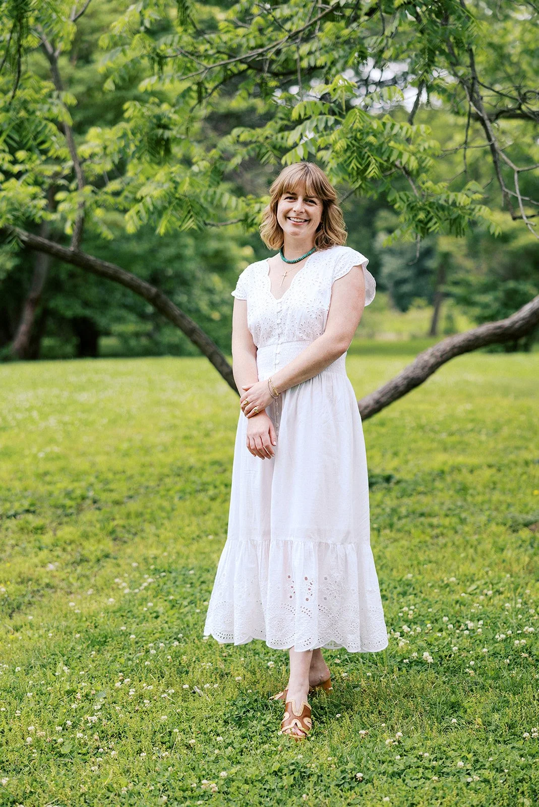 Laura Rudicle stands outdoors smiling in a white dress and blue necklace under a large tree