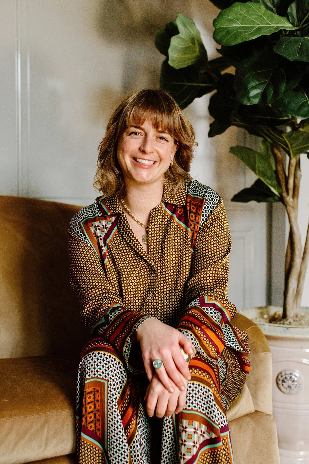 photo of Laura seated and smiling at the camera, hands overlapped with a blue ring on top, wearing a colorful patterned outfit and a gold necklace while sitting on a gold couch in front of a fiddle leaf fig tree