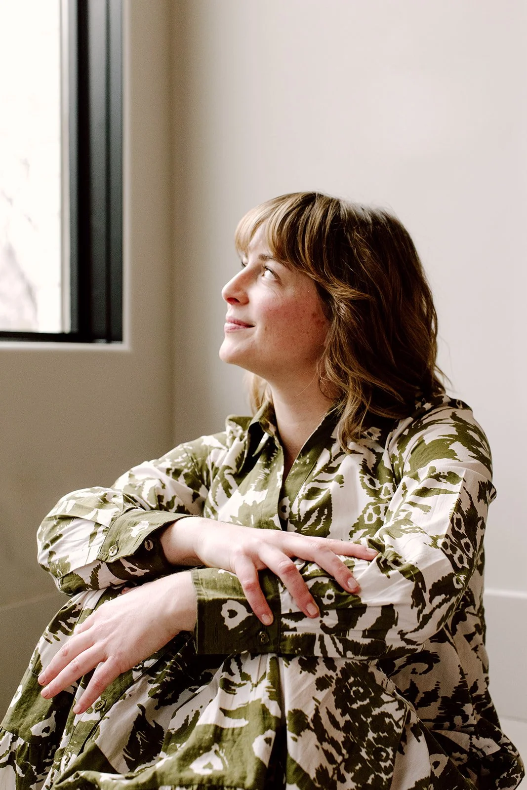 photo of Laura Rudicle sitting on the floor looking up and out the window, arms overlapped on knees, wearing a green and white patterned floral dress with a color