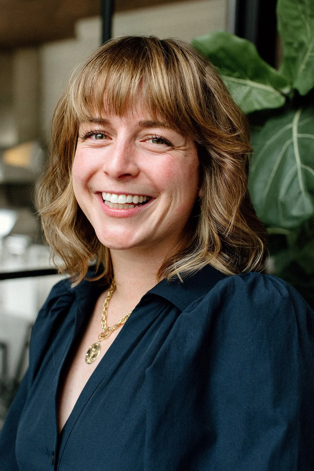 Full smile photo of Laura Rudicle indoors in dark blue dress wearing a gold necklace in front of a fiddle leaf fig tree