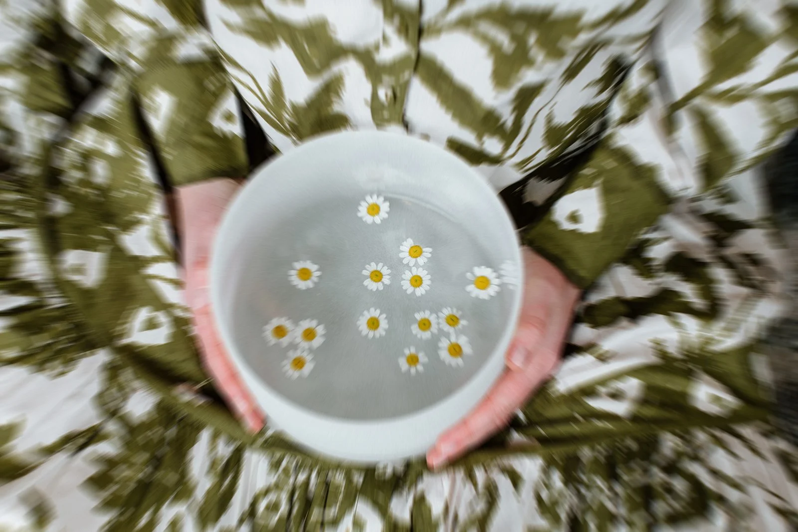 image of woman's hands holding a singing bowl in her lap on top of a green patterned dress, daisies in the bowl, and blur for motion effect