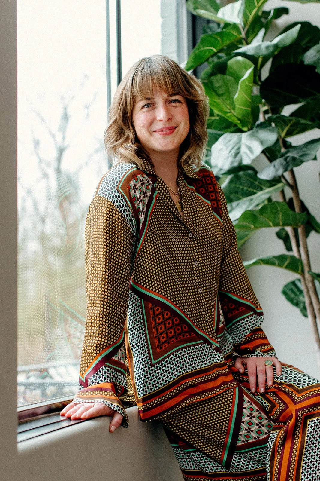 Therapist, Laura Rudicle soft smiling and setting on the ledge of a windowsill with plants in the background