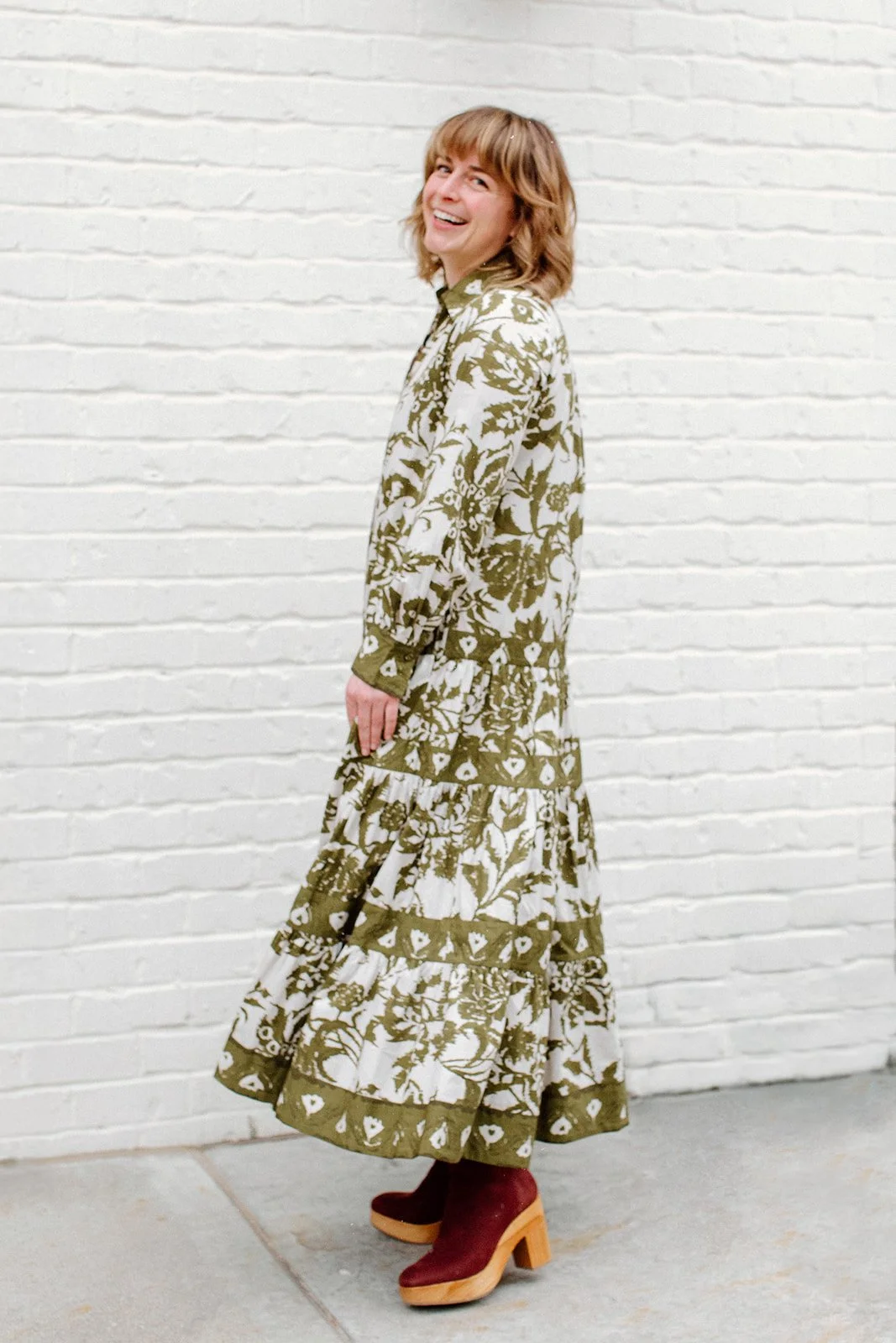 Photo of laura looking over her shoulder similing in front of a white brick wallwhile wearing a green floor-length floral dress with long sleeves and a collar.