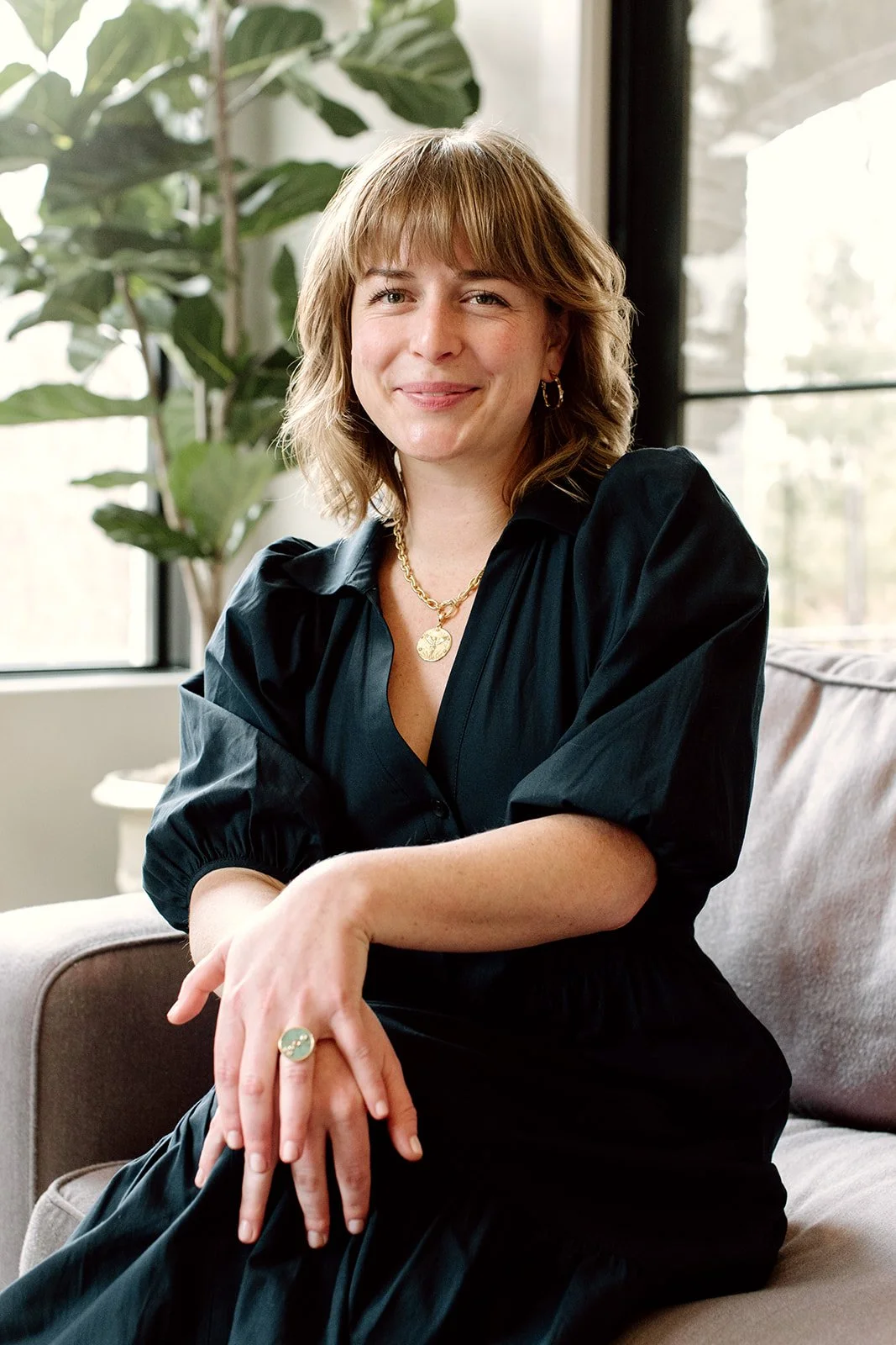 photo of Laura Rudicle soft smiling at camera seated in dark blue dress with plants in the background