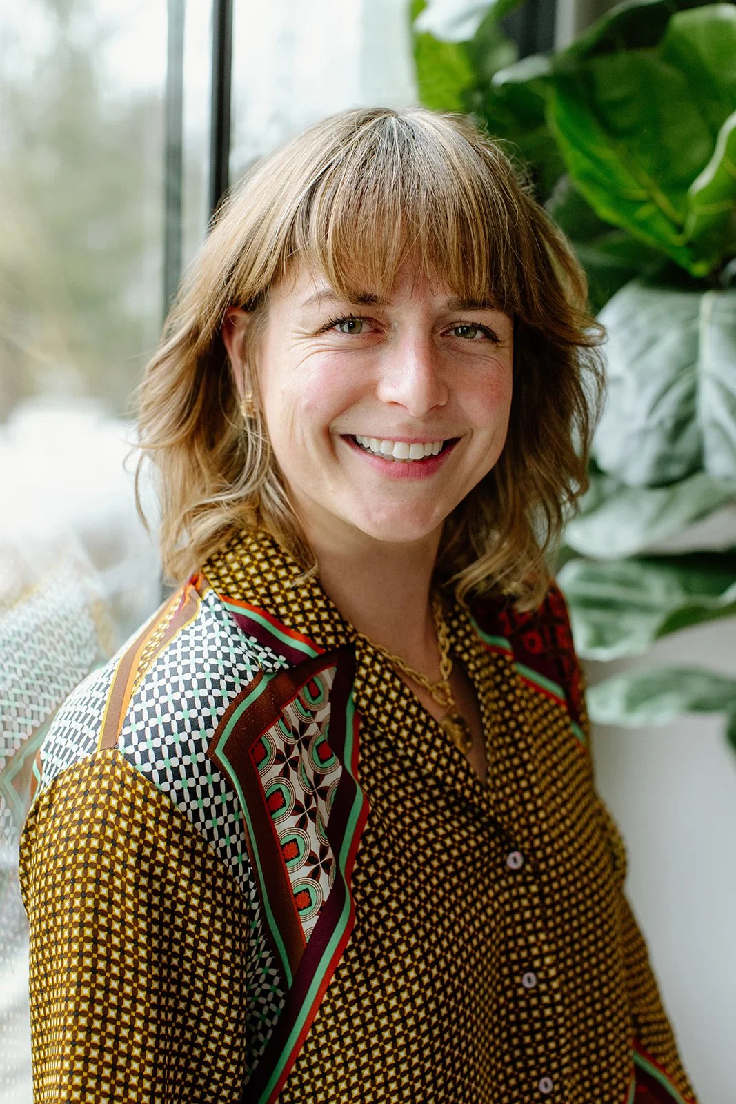 headshot of Laura Rudicle, therapist and wellness professional, smiling with teeth at camera and wearing a colorful, patterned outfit with plants in the background if the setting