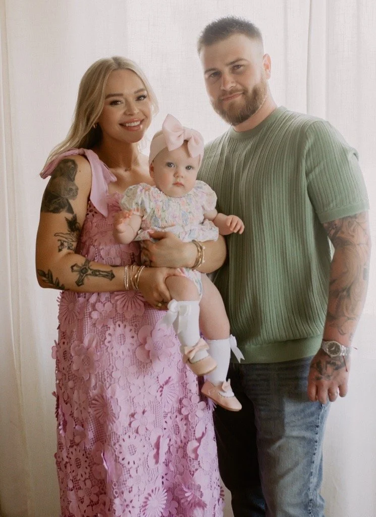 A family portrait of two adults and a baby girl. The woman has light blonde hair and tattoos on her arms, and is wearing a pink lace dress. The man has short hair and a beard, wearing a green t-shirt. The baby girl, held by the woman, wears a floral dress with a large pink bow in her hair and white socks.