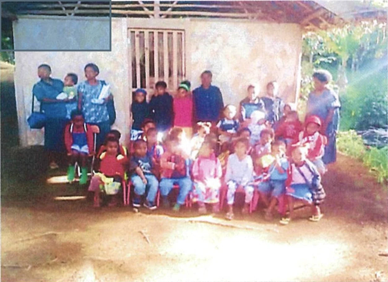 A group of children and adults gathered outdoors in front of a simple building with a wooden door and wall. Some children are sitting on pink chairs, and others are standing behind them. The scene appears to be a community or school gathering, with s