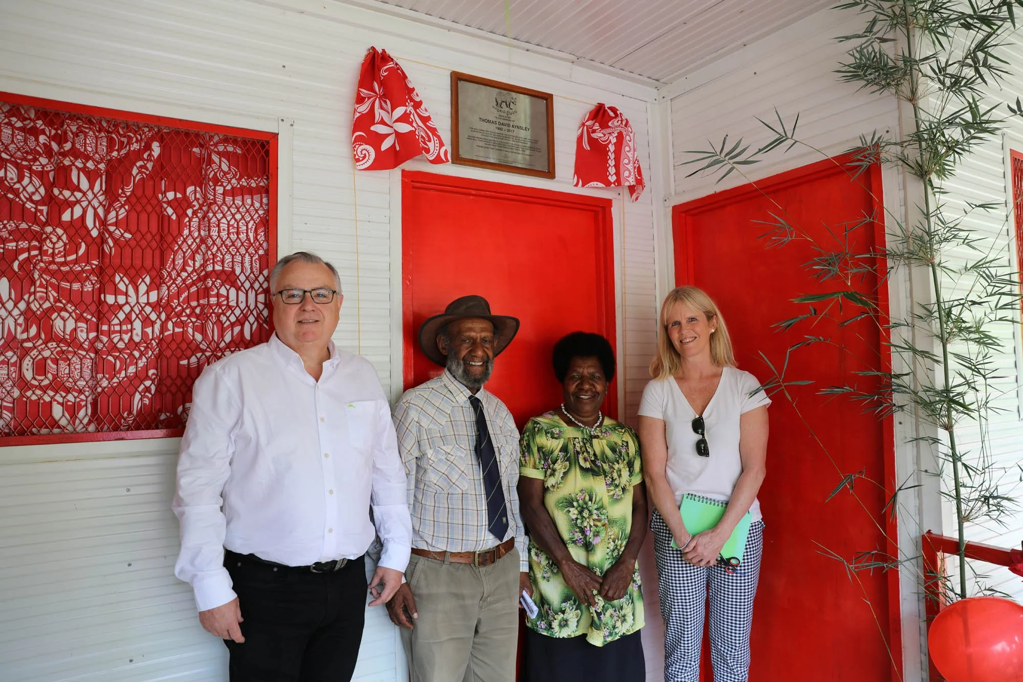 Four people standing in front of a white wall with red decorations, including two red doors and a plaque, smiling for the camera.