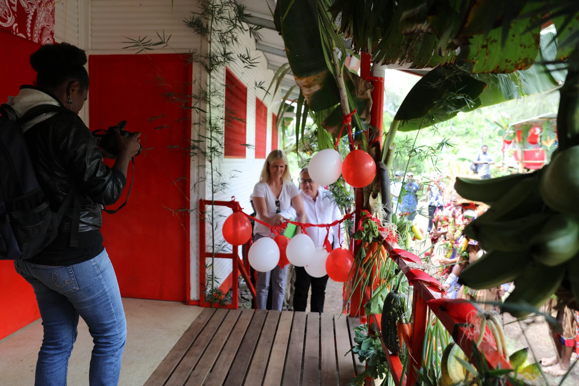 A woman is taking a photo of two people standing behind a barrier decorated with balloons and greenery. The setting appears to be a festive outdoor event with colorful decorations and a crowd in the background.