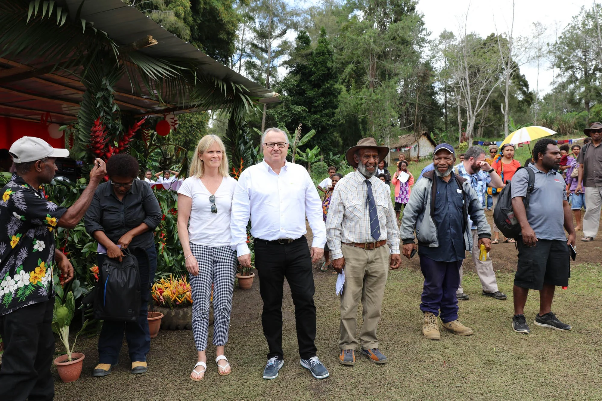 Group of people standing outdoors in a rural setting, with lush greenery and trees in the background. Some are dressed casually, while others are in more formal attire. There are potted plants and tropical decorations nearby.