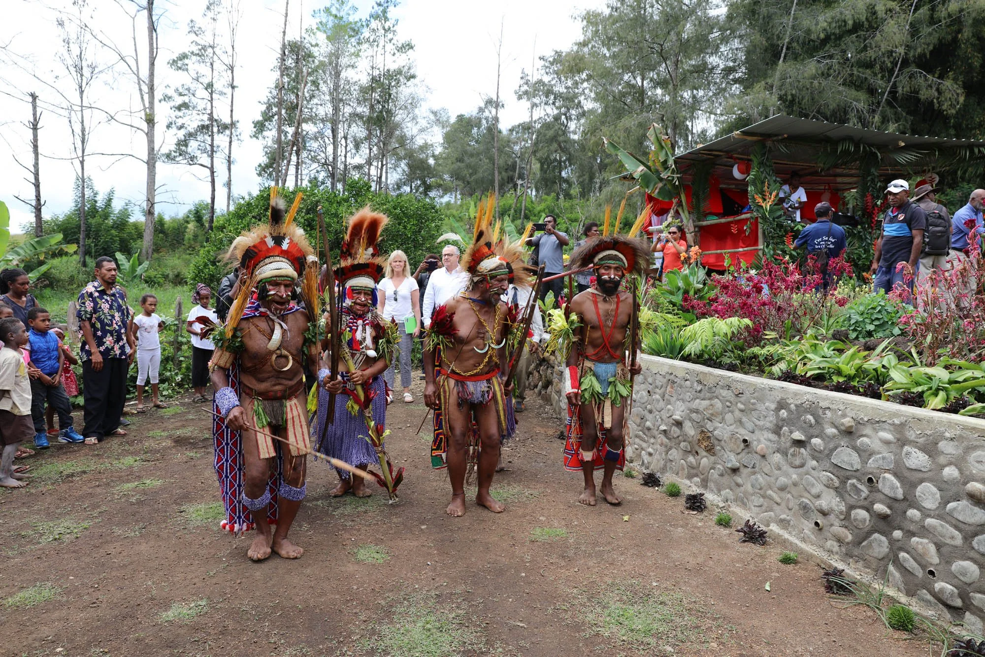 Traditional indigenous dancers in ceremonial attire performing on a dirt ground with onlookers and a platform decorated with lush plants in the background.