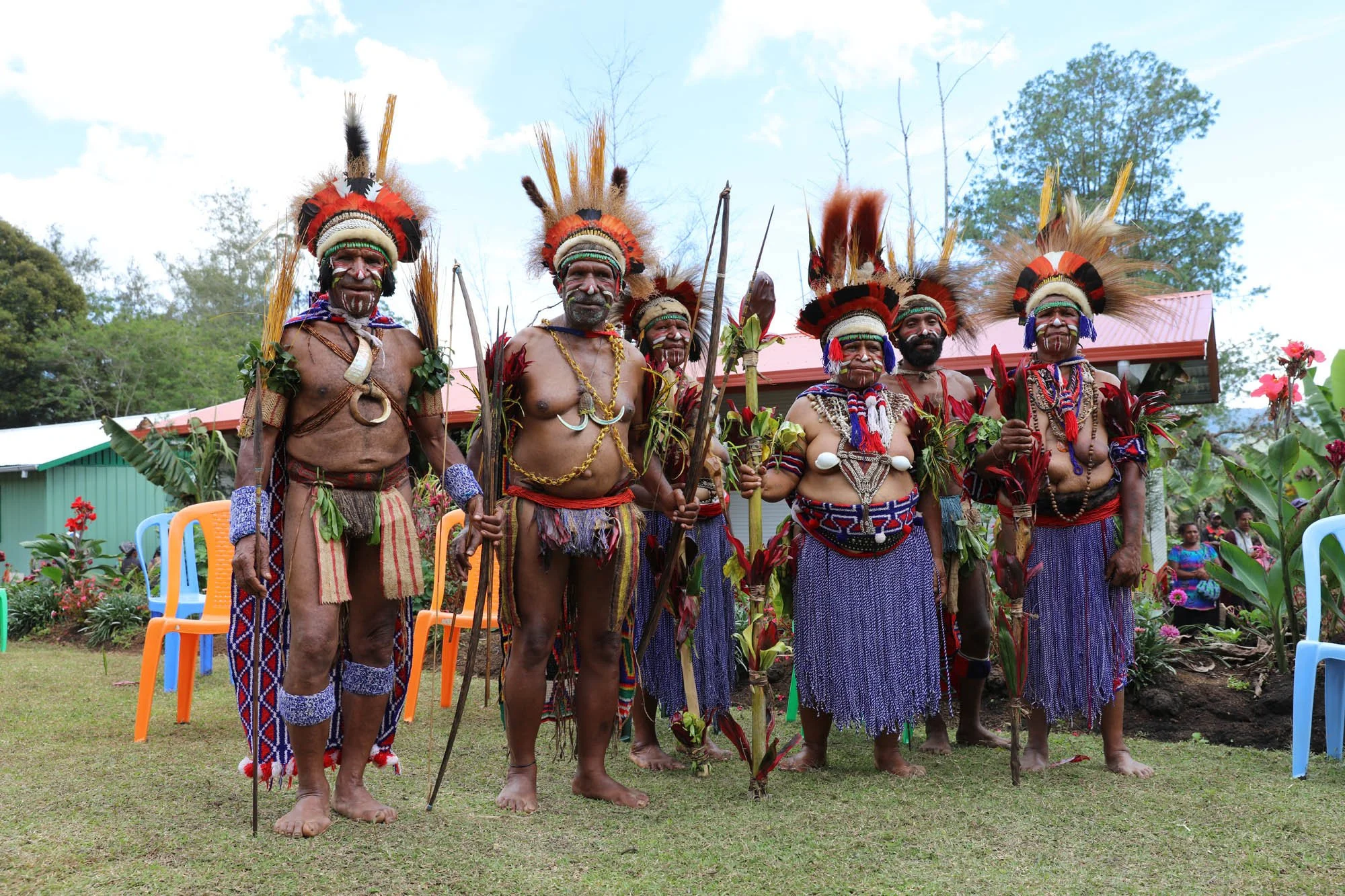 Group of indigenous men in traditional attire standing outdoors, wearing feathered headdresses, body paint, and holding sticks, with some holding flowers.