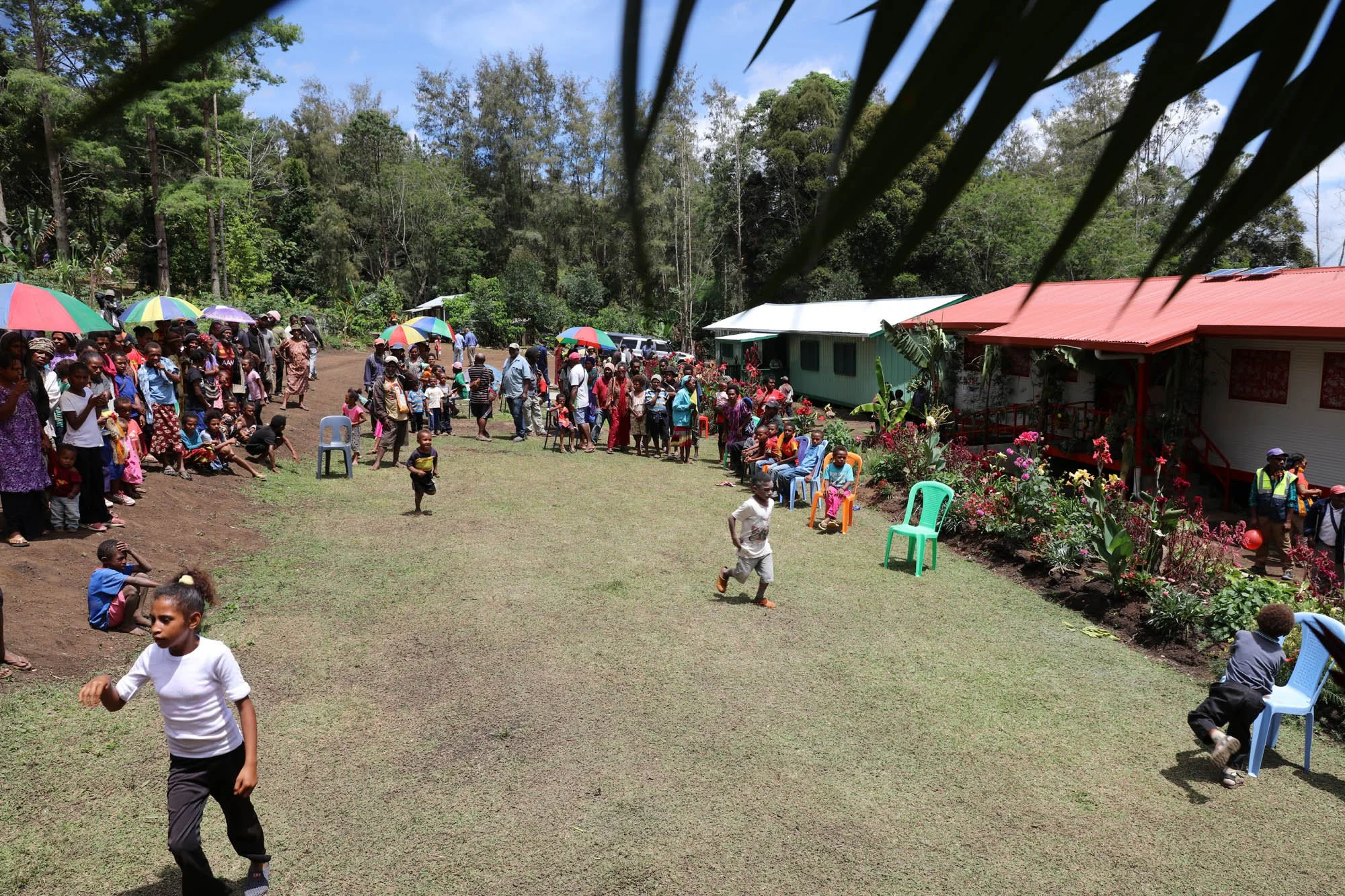 Crowd of people, including children, gathered outdoors on a grassy area with some sitting on chairs and others standing, under umbrellas in a rural setting with trees and houses in the background.