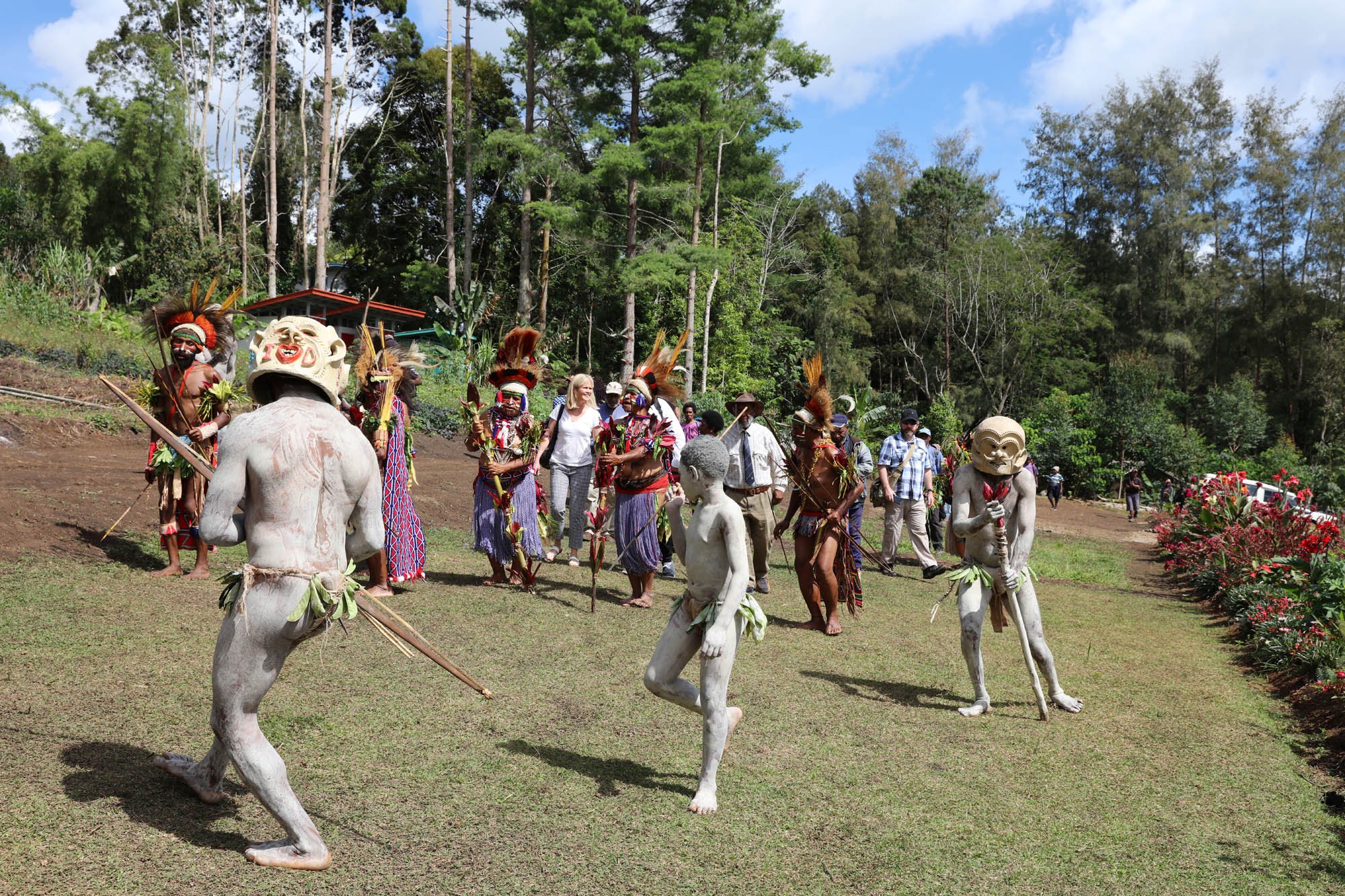 A cultural scene featuring individuals in traditional tribal costumes with masks and body paint, performing a dance outdoors on green grass with trees and a blue sky in the background.