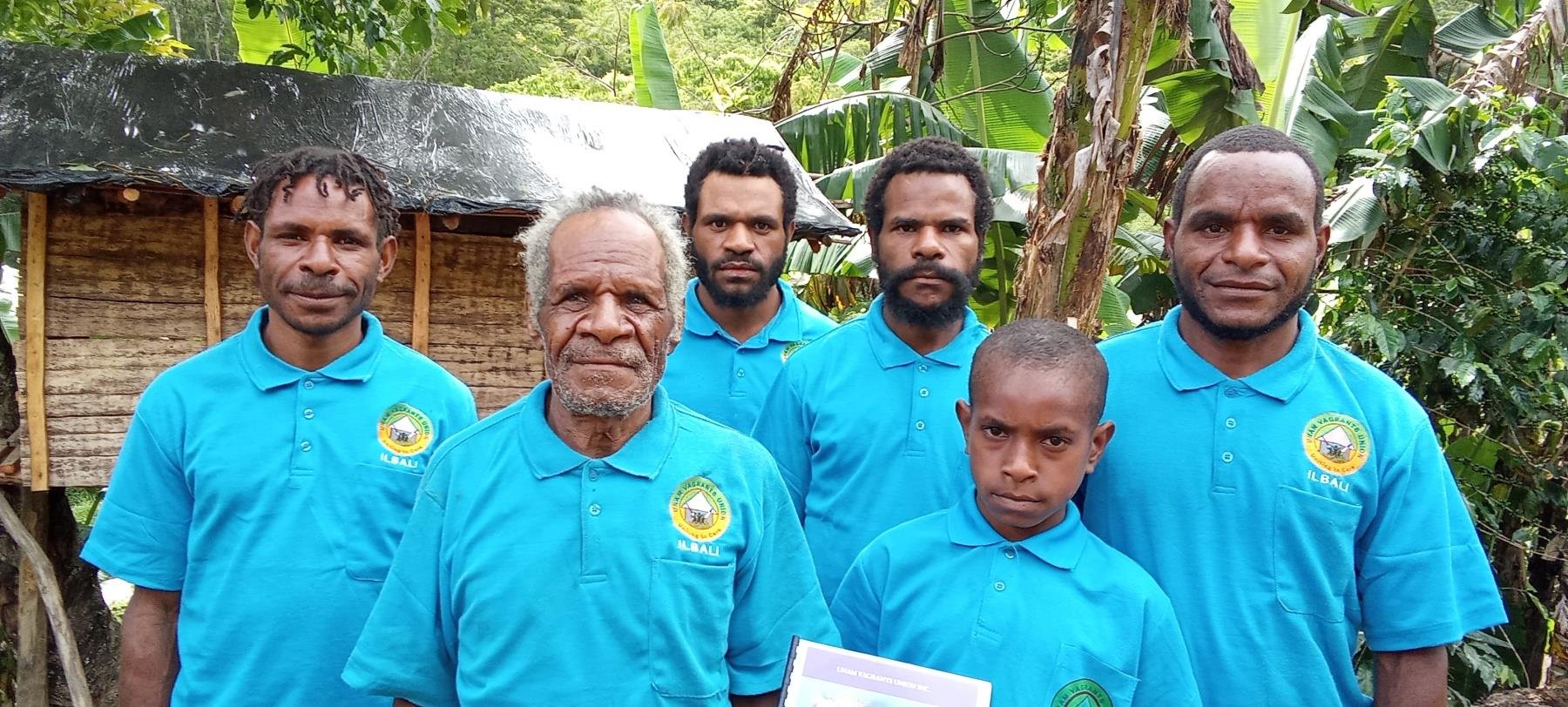 Group of six men and one boy standing outdoors in front of banana trees and a small wooden hut, all wearing blue shirts with a logo on the left chest.