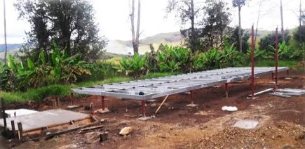 Construction site with a concrete slab foundation on dirt, supported by metal poles, in a rural area with trees and green vegetation.