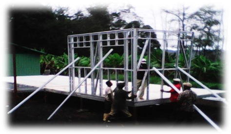 Construction workers assembling a metal frame structure outdoors on a platform with trees in the background.