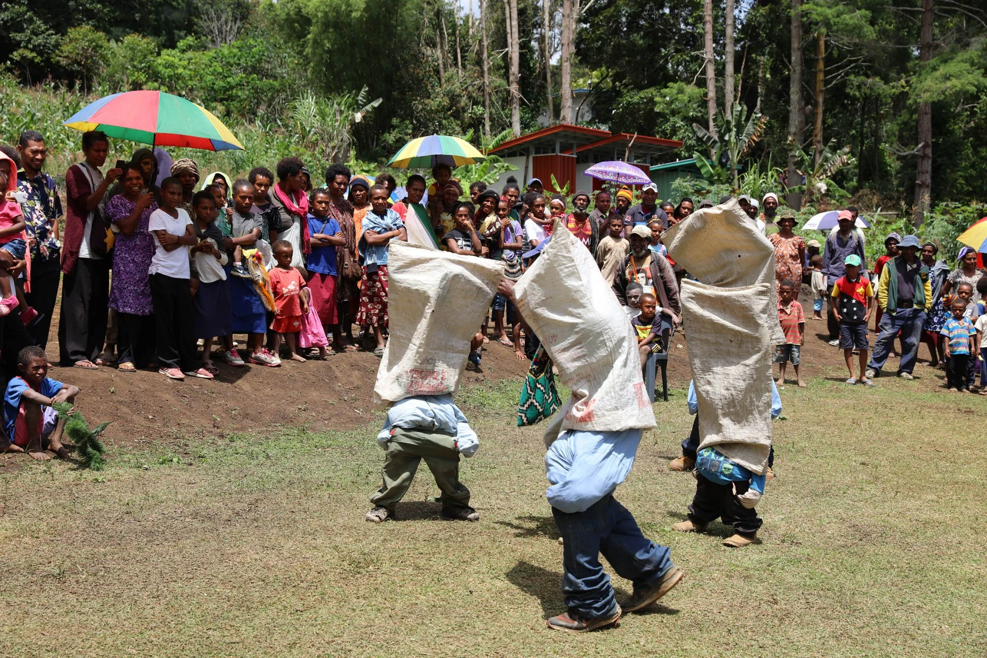 Children and adults gathered outdoors, some holding umbrellas, watching a performance of children with sacks jumping on the ground.