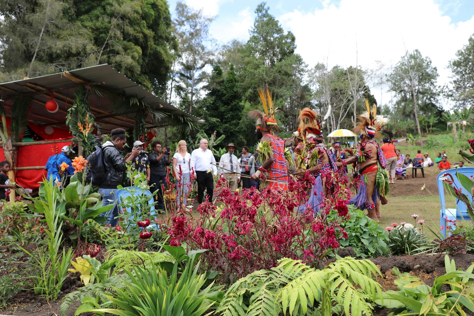 Traditional indigenous dance performance outdoors with dancers in colorful costumes and feathered headpieces, surrounded by lush green plants, spectators, and a small decorated stage in a rural setting.