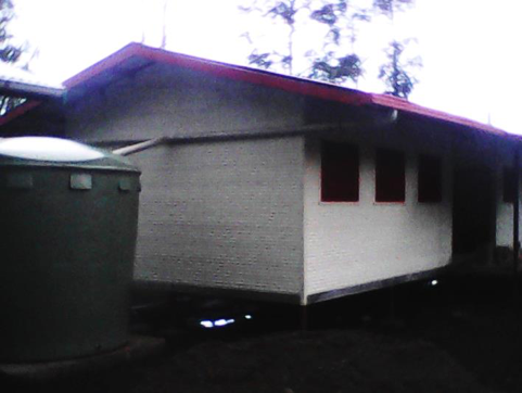 A small, elevated house with a red roof, three red-trimmed windows, and a water tank nearby, under a partly cloudy sky.