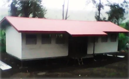 A small house with a red metal roof and white walls, situated outdoors with trees in the background.