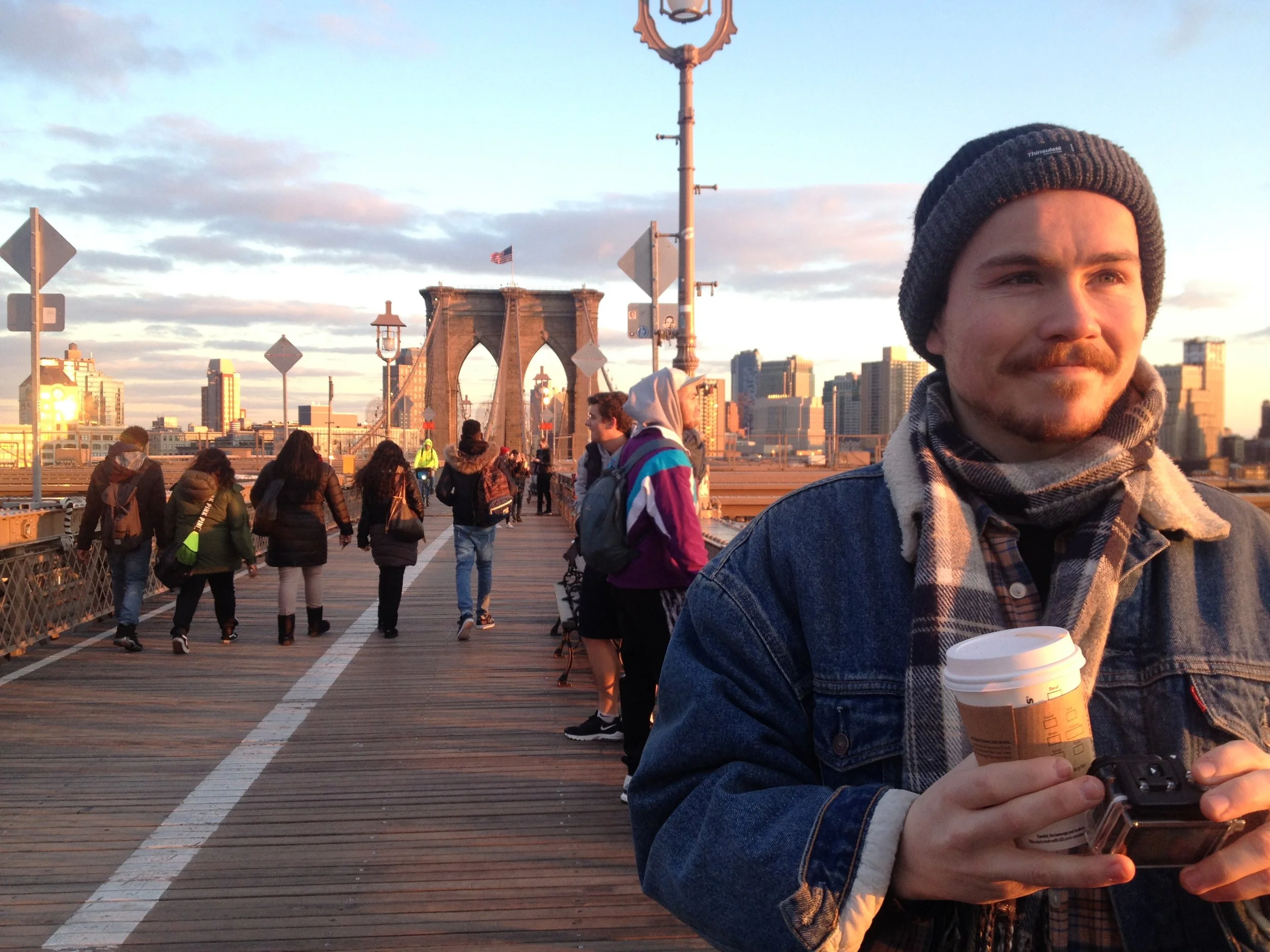A man with a beard and mustache wearing a beanie, scarf, and denim jacket holding a coffee cup and camera in front of Brooklyn Bridge at sunset with pedestrians walking on the bridge walkway.