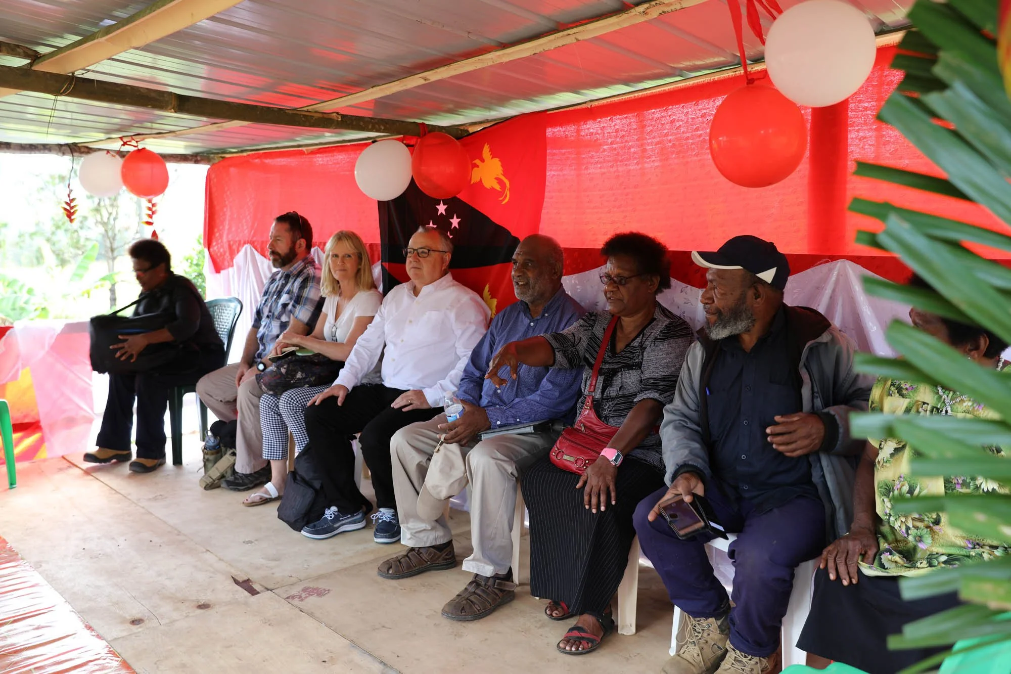 Group of diverse people sitting together in a decorated outdoor setting with red and white balloons, a red backdrop, and tropical plants.