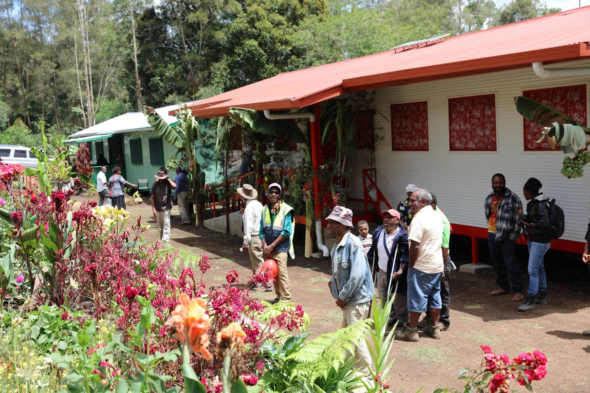 A group of people standing outside in a garden area with flowering plants, next to a building with red windows and a red roof.