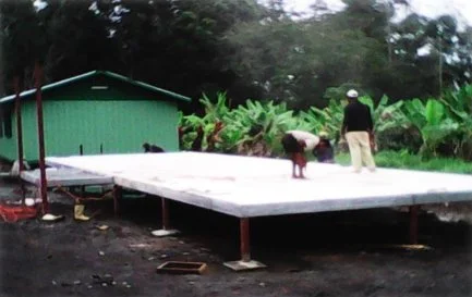 Two people working on a construction project on a raised platform outdoors, with greenery and a small building in the background.