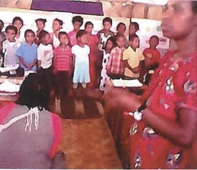 Group of children standing in a classroom, with a woman speaking or leading an activity at the front.