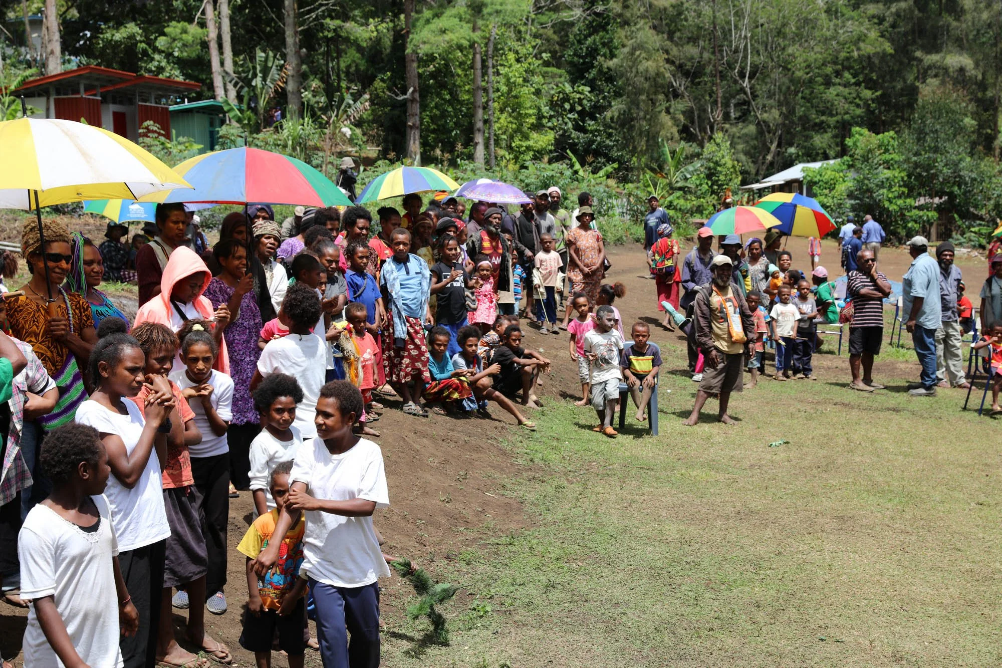 A large group of people, including children and adults, gathered outdoors on a grassy area with trees in the background, holding colorful umbrellas.