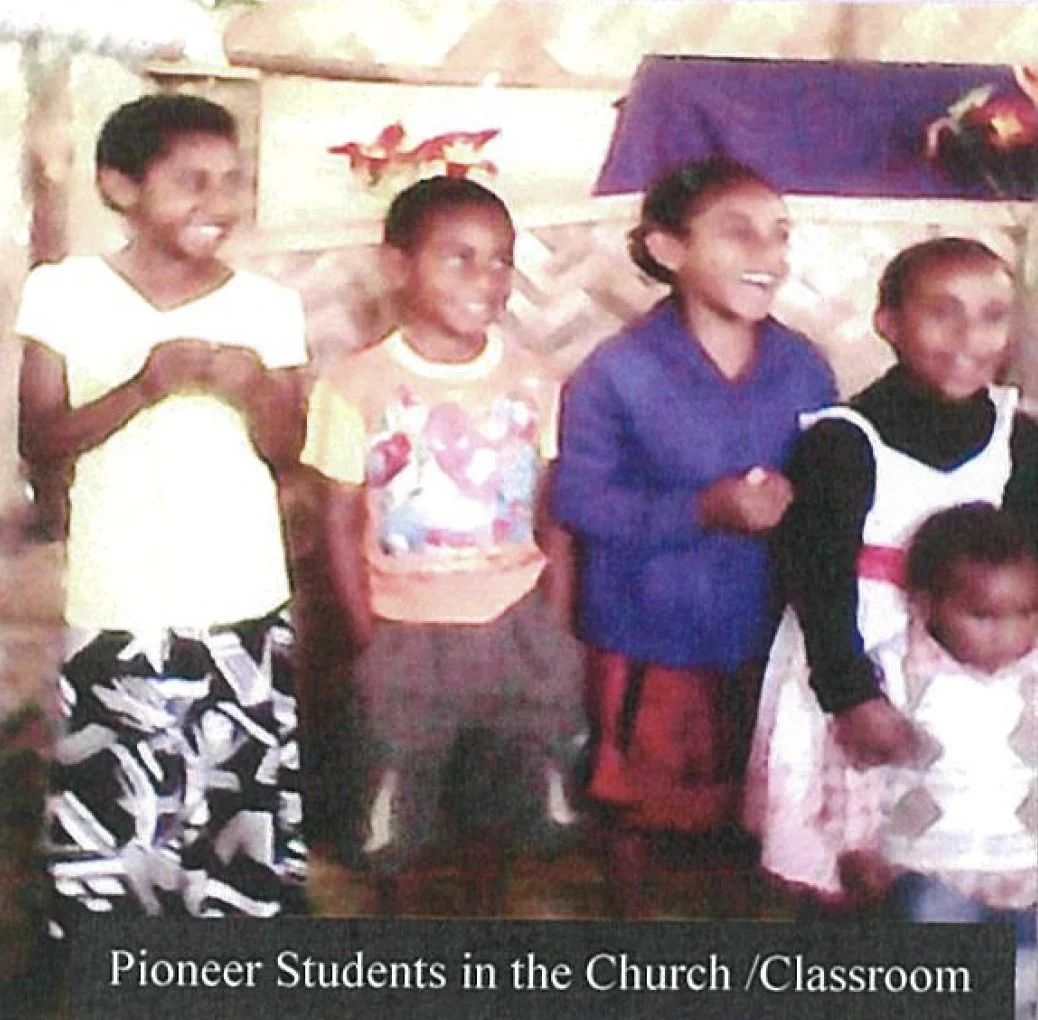 Group of five young children, four girls and one boy, standing inside a room, smiling, with a girl holding a young child. The background has a wooden wall with decorations and a table covered with a cloth. Caption reads, 'Pioneer Students in the Chur
