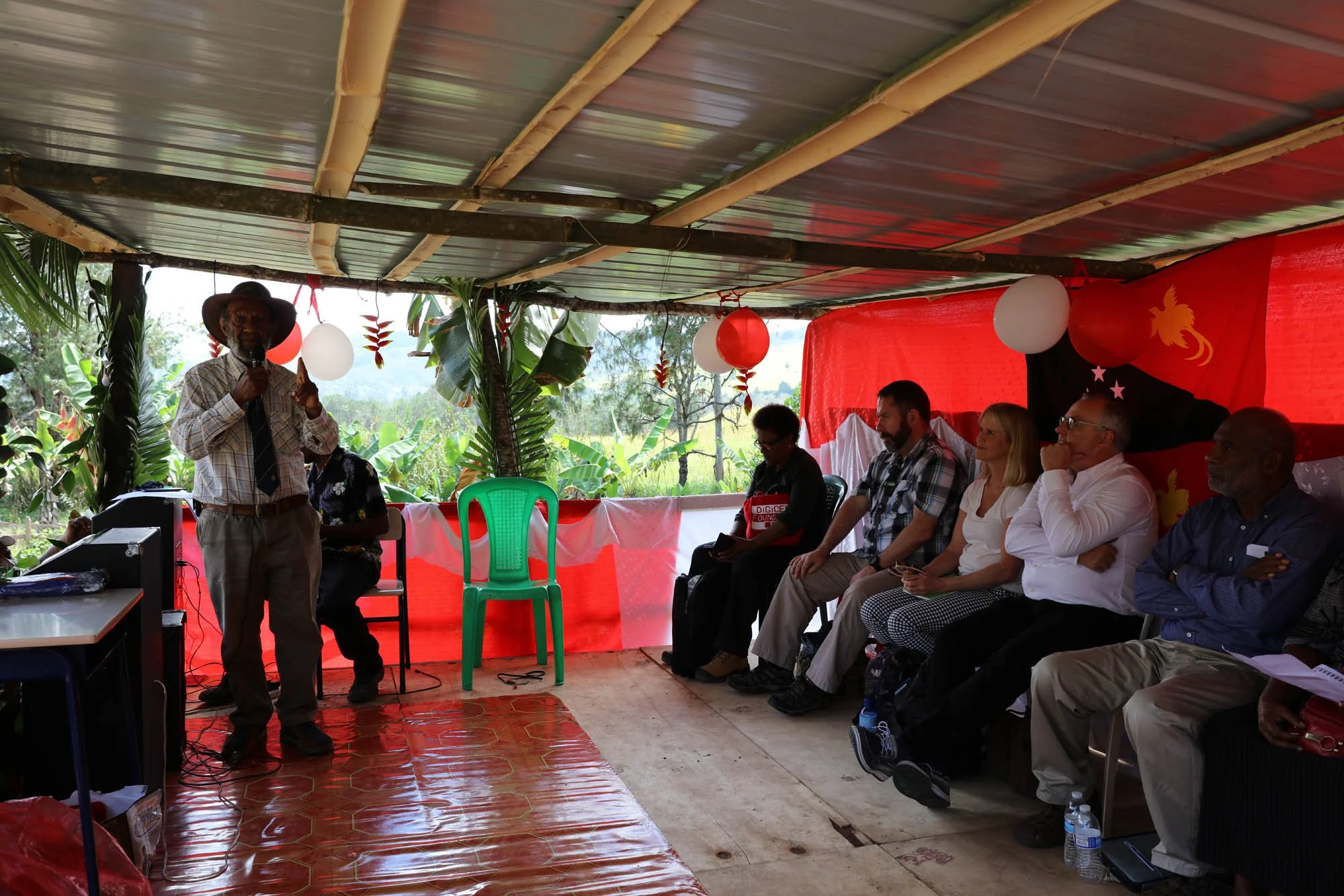 A man in a plaid shirt and tie giving a speech to a group of people seated under a decorated shelter with red and white balloons and greenery background.