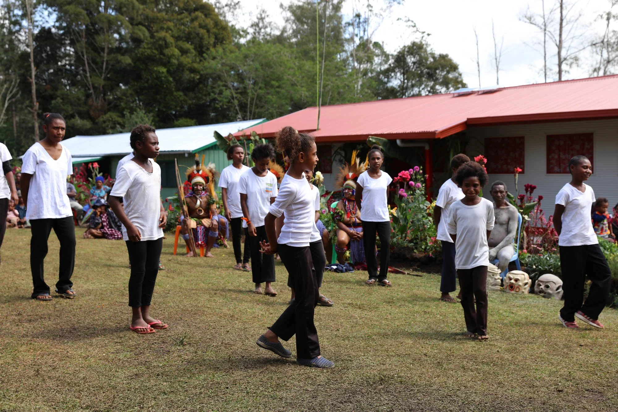 Children and adults performing traditional dance outdoors with spectators and indigenous cultural attire in the background.